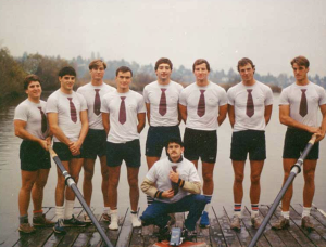 A group of young men stand in white shirts with red tikes and black shorts holding 2 oars on a dock with a lake in the background. One young man kneels in front of them.